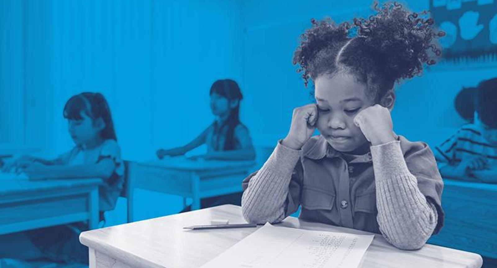 A child at their classroom desk in grayscale foreground looking at their problem sheet.