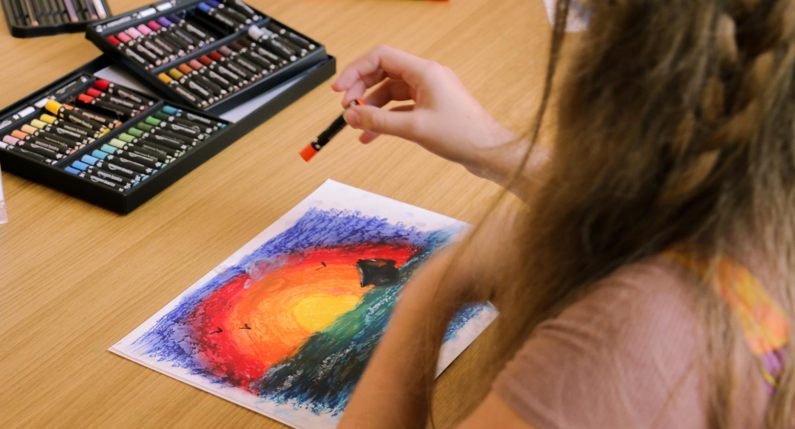 A student draws a sunset with oil pastels in the Contemplative Commons. Other art supplies are present on the table, such as markers, colored pencils, and more oil pastels. 