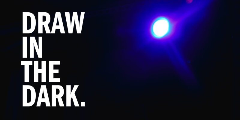 An image of a blue light shining over a dark room from a floor perspective. Bold, white text to the left of the light says "DRAW IN THE DARK." in all capital letters. 