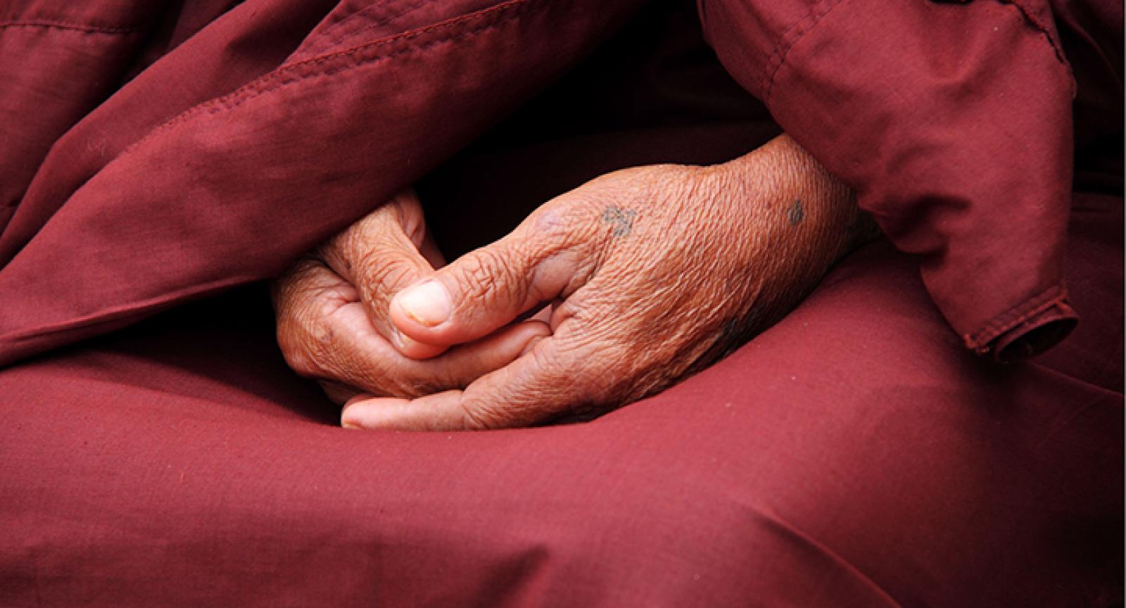 Image of a monk’s hand joined in their lap with the red robes behind them