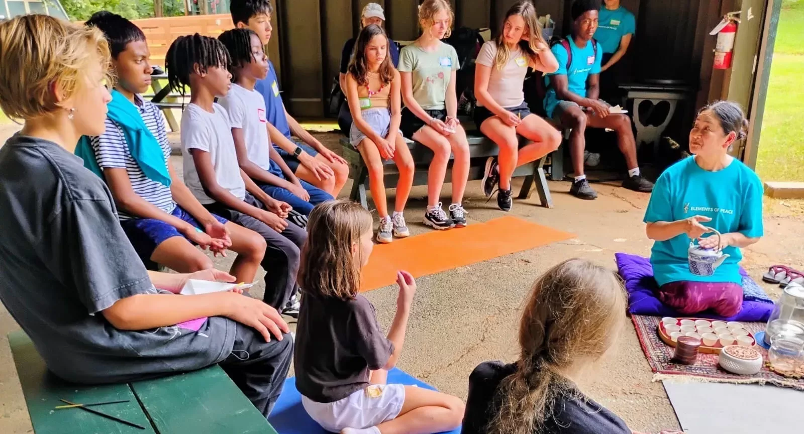 Children sit on benches around a teacher pouring tea