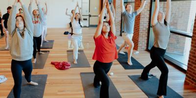 Students practice Ashtanga Yoga, reaching their arms up towards the ceiling in a room with wooden floors