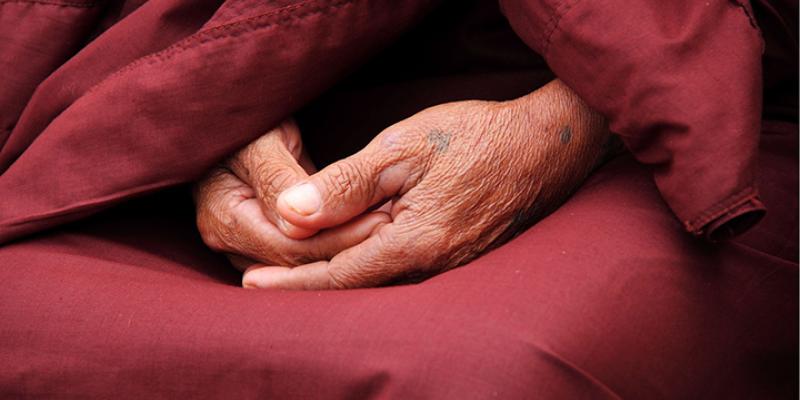 Image of a monk’s hand joined in their lap with the red robes behind them