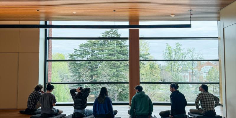 a group of students sits on a wooden floor in the Contemplative Commons and meditates, overlooking the foliage below from the floor to ceiling window