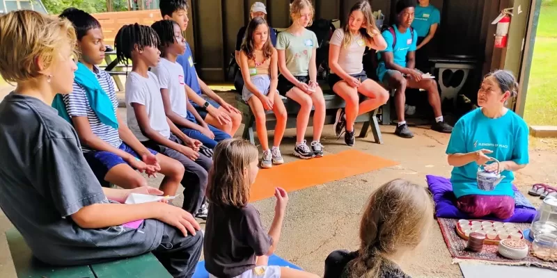 Children sit on benches around a teacher pouring tea