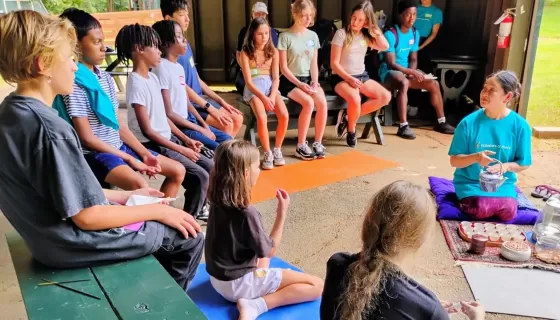 Children sit on benches around a teacher pouring tea