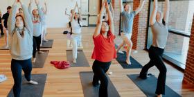Students practice Ashtanga Yoga, reaching their arms up towards the ceiling in a room with wooden floors