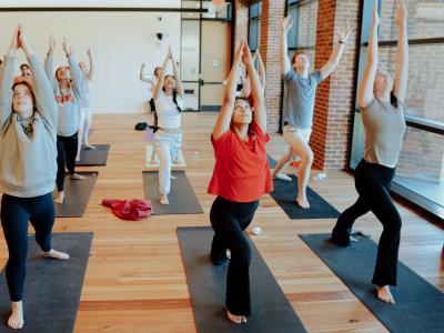 Students practice Ashtanga Yoga, reaching their arms up towards the ceiling in a room with wooden floors