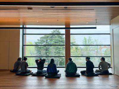 a group of students sits on a wooden floor in the Contemplative Commons and meditates, overlooking the foliage below from the floor to ceiling window