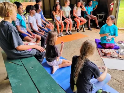 Children sit on benches around a teacher pouring tea