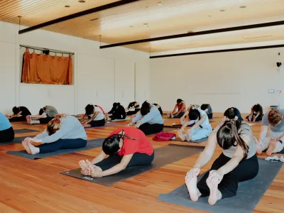 A group of students practicing yoga. They sit with their legs straight and reach towards their toes. 